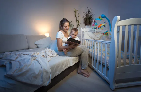 Madre joven sentada en la cuna del bebé y leyendo un libro —  Fotos de Stock