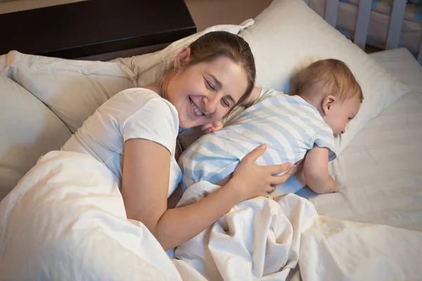 Noche de feliz madre sonriente acostada en la cama con su pequeño —  Fotos de Stock