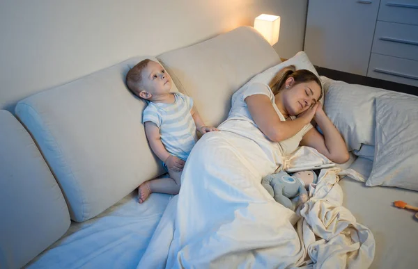 Sleepless baby boy waking up his mother sleeping in bed — Stock Photo, Image