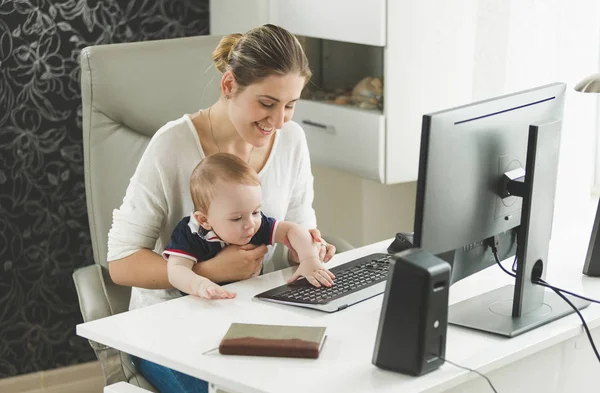 Mujer sonriente trabajando en casa y sosteniendo a su hijo en el regazo —  Fotos de Stock