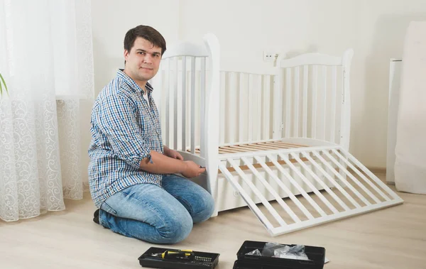 Young man assembling white wooden crib in nursery