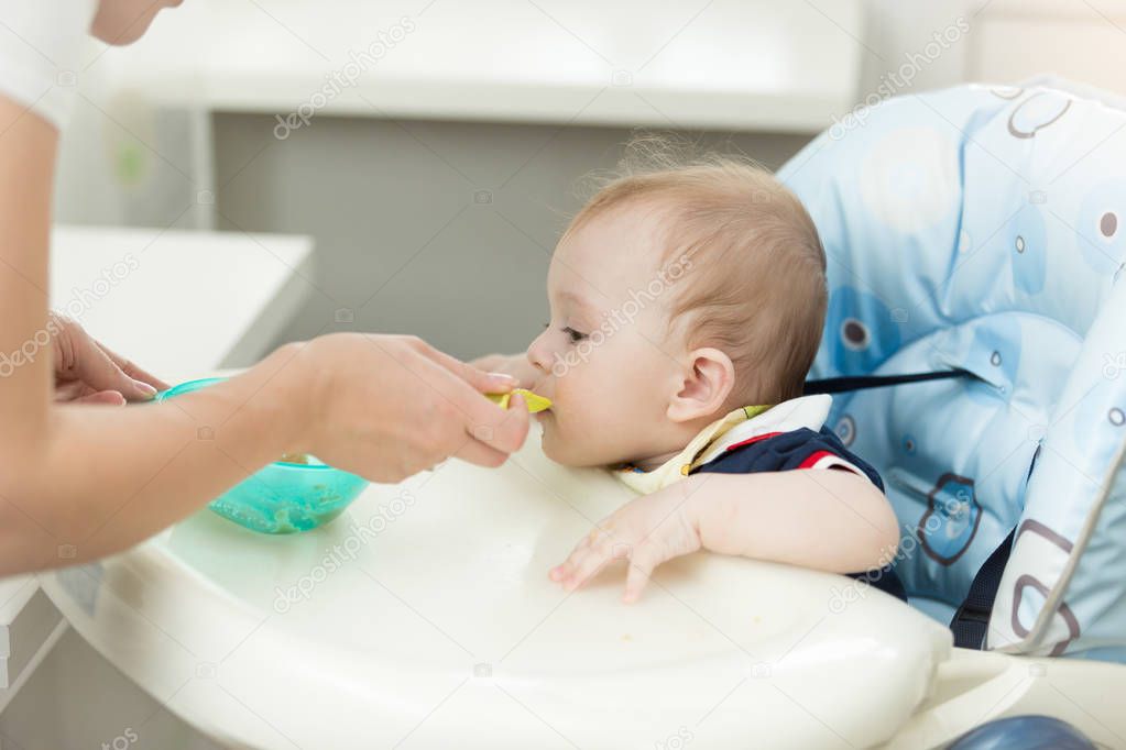 Baby boy sitting in highchair at kitchen and eating from spoon