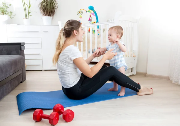 Hermosa madre jugando con su bebé en la alfombra de fitness en casa — Foto de Stock