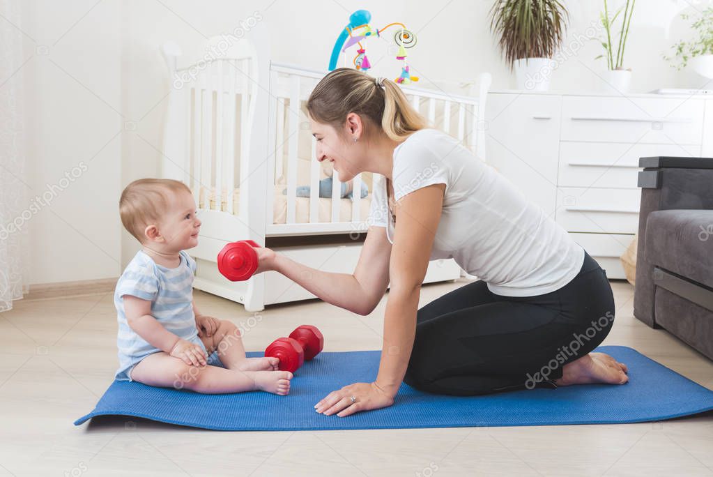 Young mother doing fitness and giving dumbbells to her baby. Con