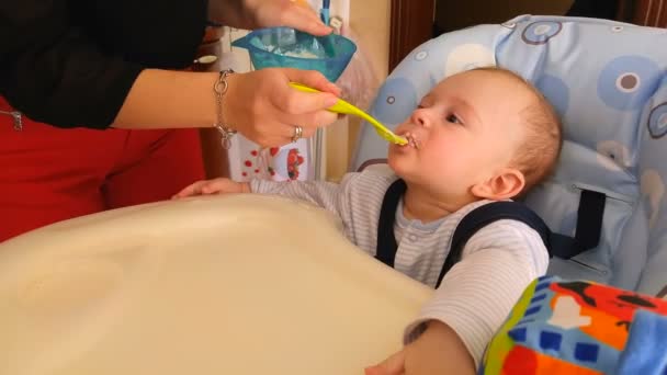 Молодая мать кормит симпатичного маленького сына сидя в детском кресле на кухне — стоковое видео