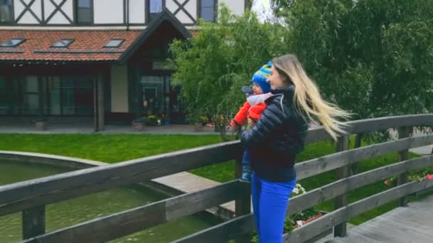 Feliz joven madre de pie con su bebé hijo puente de madera en el frío día ventoso — Vídeos de Stock