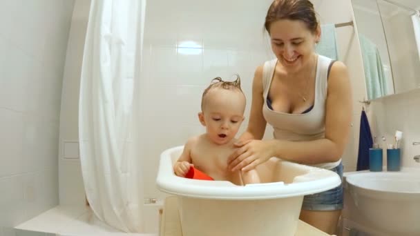 プラスチック製の小さな浴槽で彼女の赤ちゃんを洗う若い美しい母親のスローモーション — ストック動画