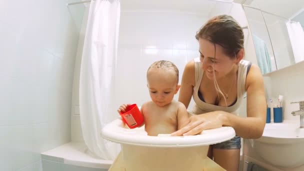 Прекрасная любящая мать целует своего маленького сына, купающегося в пластиковой ванне — стоковое видео