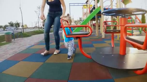 Bonito menino sorridente andando com a jovem mãe no parque infantil no carrossel — Vídeo de Stock