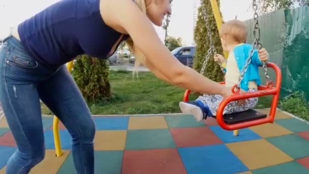 Zeitlupe zeigt junge Mutter, die ihren kleinen Sohn auf Spielplatz schaukelt — Stockvideo