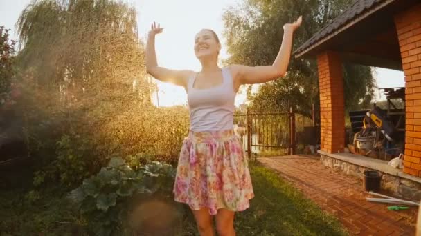 Медленное видео, где женщина танцует на закате под водой из садового разбрызгивателя — стоковое видео