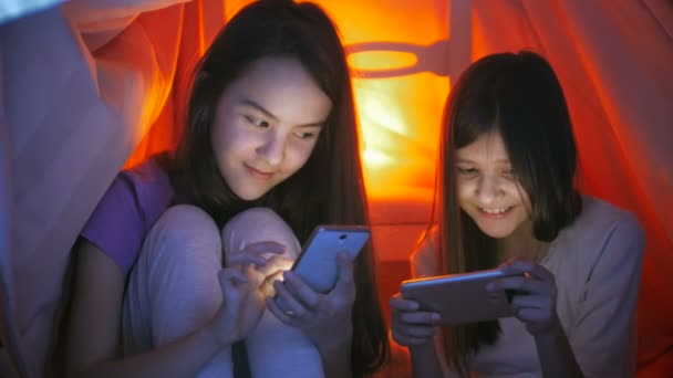 4k vídeo de duas meninas sorridentes bonitos sentados sob cobertor à noite e usando telefones celulares — Vídeo de Stock