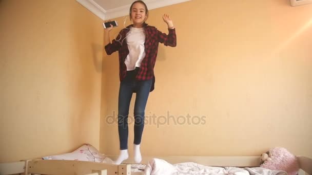 Vídeo em câmera lenta de uma adolescente feliz ouvindo música com fones de ouvido e pulando no colchão no quarto — Vídeo de Stock