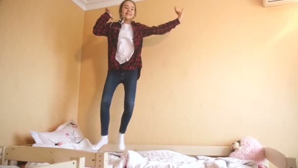 Медленное движение веселой девочки-подростка слушает музыку по мобильному телефону и прыгает на кровать — стоковое видео