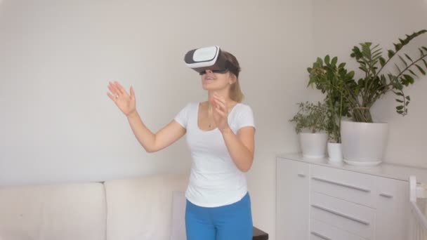 Удивительная молодая женщина в VR гарнитуре ходит в комнату и ловит предметы перед ней. Съемка на 4k — стоковое видео