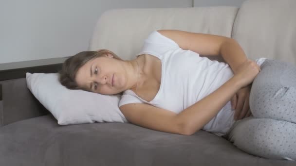 Молодая женщина страдает от боли в животе лежа на диване в гостиной — стоковое видео