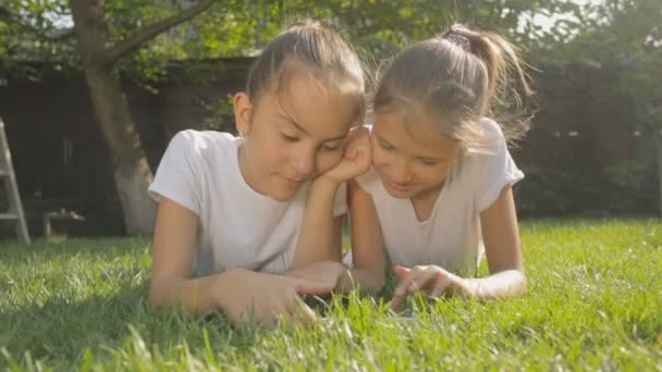 Zwei lächelnde Mädchen, die sich im Gras entspannen und auf einem digitalen Tablet spielen — Stockvideo