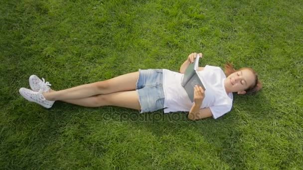 Вид сверху на красивую девочку-подростка, лежащую на траве и читающую книгу — стоковое видео