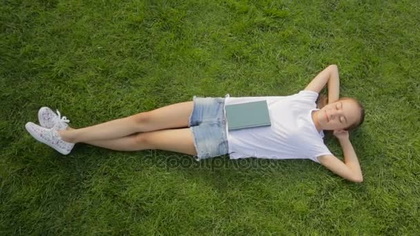 Belle fille yong se détendre sur l'herbe verte au parc avec livre — Video