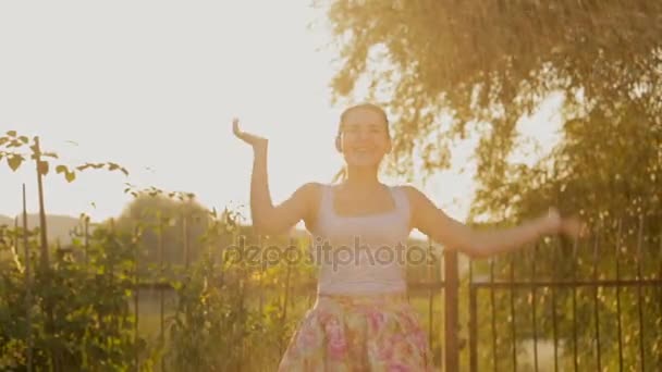 Feliz joven mujer bailando bajo el agua del rociador de jardín al atardecer — Vídeos de Stock