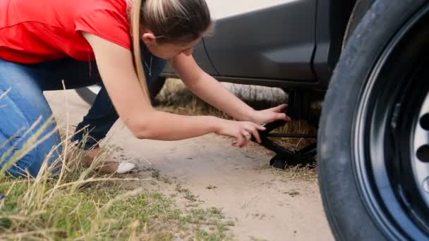 Young woman using jack-screw to lift a car before changing flat tire on dirt road at countryside — Stock Video