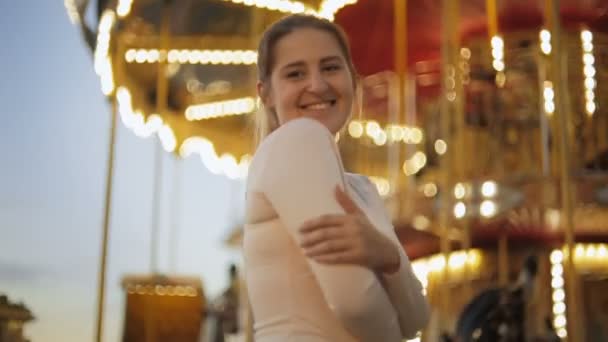 Портрет красивой улыбающейся женщины, стоящей на освещенной карусели в парке развлечений — стоковое видео