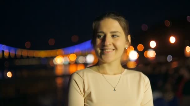 Dolly disparo de mujer joven elegante posando en el puente iluminado sobre el río por la noche — Vídeos de Stock