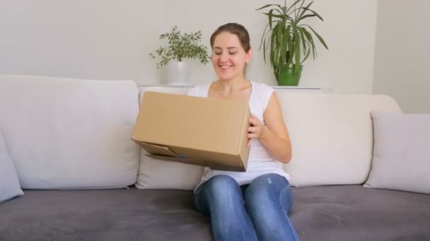 Mujer joven y feliz sentada en el sofá en la sala de estar y tomando vestido de la caja que compró en la tienda en línea — Vídeo de stock