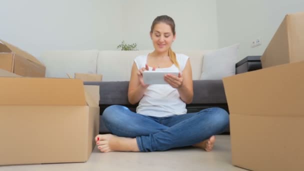 Dolly shot of happy young woman packing things in cardboard boxes and using digital tablet — Stock Video