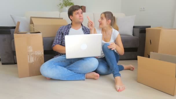 Casal jovem feliz desempacotando caixas em sua nova casa e escolhendo móveis no laptop — Vídeo de Stock