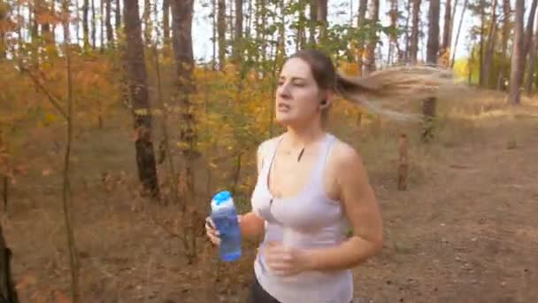 Imágenes de 4K de una hermosa joven corriendo en el bosque y bebiendo agua de la botella — Vídeos de Stock