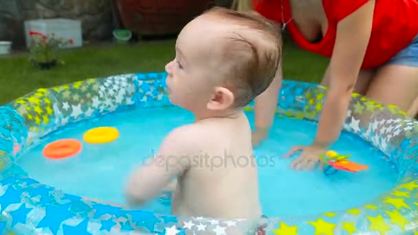 Милый мальчик плавает и играет с игрушками в надувном бассейне на траве в саду — стоковое видео