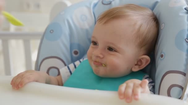 Fechar-se de jovem mãe alimentando bonito sorrindo bebê menino vestindo babador e sentado em cadeira alta — Vídeo de Stock