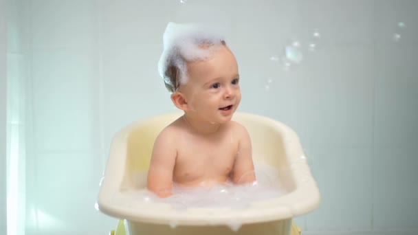 Filmagem em câmera lenta de bebê adorável tomando banho e olhando para bolhas de sabão coloridas — Vídeo de Stock