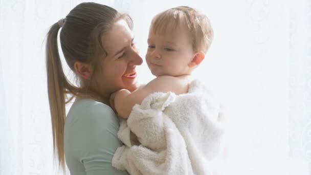 Медленная съемка красивой улыбающейся матери, обнимающей и разговаривающей со своим ребенком у большого окна — стоковое видео