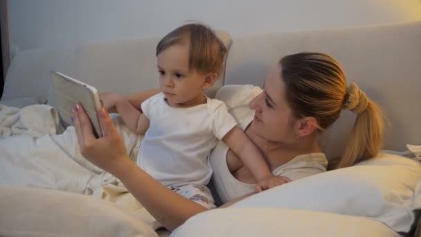 Cute baby boy lying in bed with mother and watching cartoons on digital tablet — Stock Video