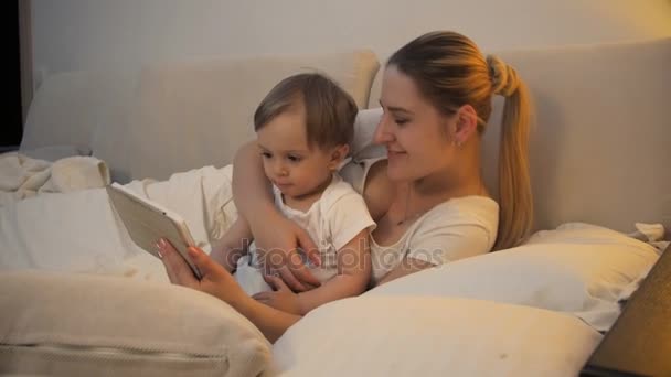 Młoda matka leży z jej synkiem na łóżku z cyfrowego tabletu — Wideo stockowe