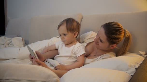 4k vídeo de bonito sorrindo bebê menino assistindo desenhos animados no tablet com a mãe na cama — Vídeo de Stock