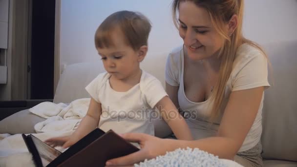 Mère avec bébé fils regardant à travers l'album photo de famille dans le lit la nuit — Video