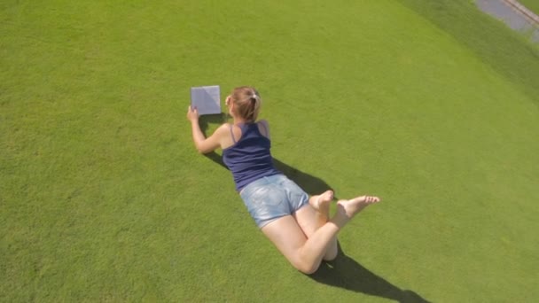 Вид сверху на молодую студентку, лежащую на траве в парке и читающую книгу — стоковое видео