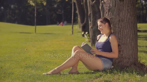 Bella donna sorridente che si rilassa sotto l'albero al parco e tiene un libro — Video Stock