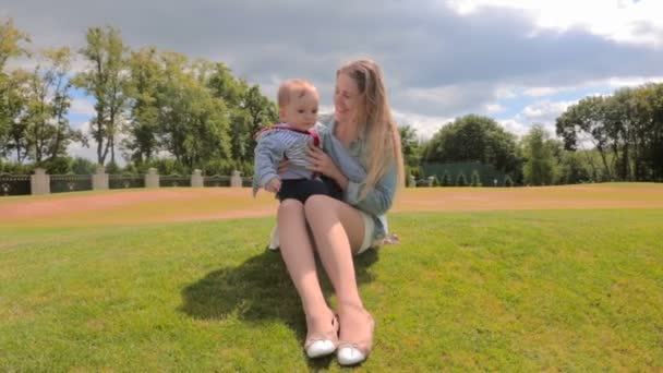 Счастливая улыбающаяся мать отдыхает со своим маленьким мальчиком на траве в парке — стоковое видео