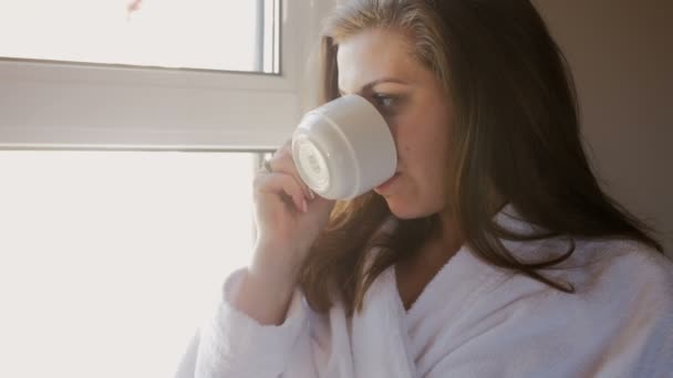 4 k 特写视频，美丽的年轻女子，穿着浴袍喝咖啡在窗口 — 图库视频影像