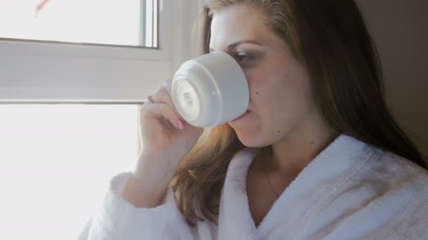 Primer plano de vídeo de una joven en albornoz bebiendo café y mirando por la ventana — Vídeo de stock