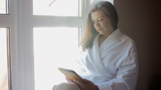 4k видео красивой молодой женщины в халате, сидящей на подоконнике и использующей цифровой планшет — стоковое видео