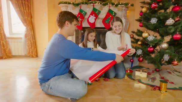Familia feliz con niña envolviendo regalos de Navidad en papel de regalo rojo — Vídeo de stock
