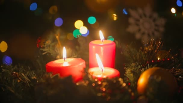 Primo piano della corona natalizia con tre candele rosse accese. Luci sfocate luminose colorate su sfondo — Video Stock