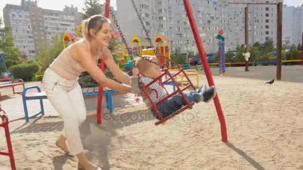 Feliz jovem mãe balançando seu bebê filho em balanços no playground — Vídeo de Stock