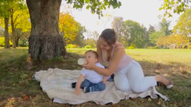 Güzel genç anne bebek oğlu şişe Park'ta ağacın altında besleme