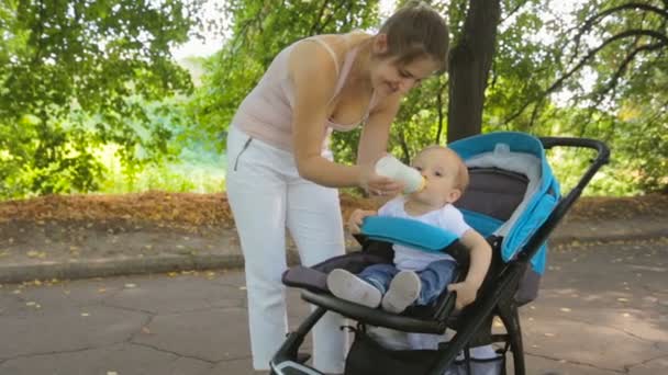 Hermosa mujer alimentando a su hijo del biberón en cochecito en el parque — Vídeos de Stock
